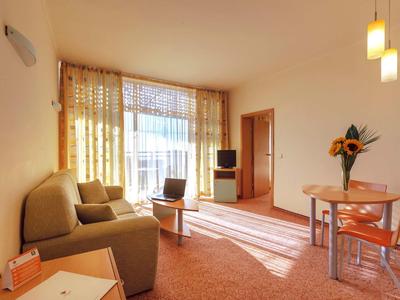 Světý obývací pokoj s pohovkou, stolem, židlemi, televizí a velkými okny se závěsy.