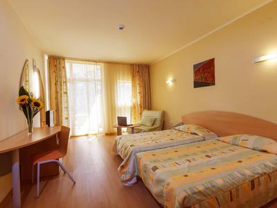 Světle hotelový pokoj se dvěma jednolůžky, psacím stolem, křeslem a velkým oknem se závěsy.