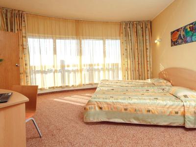 Světý hotelový pokoj s manželskou postelí, psacím stolem, židlí a velkými okny s závěsy.