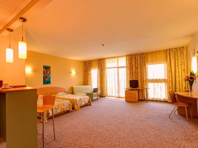 Světý hotelový pokoj s manželskou postelí, posezením, psacím stolem a balkonem se závěsy.