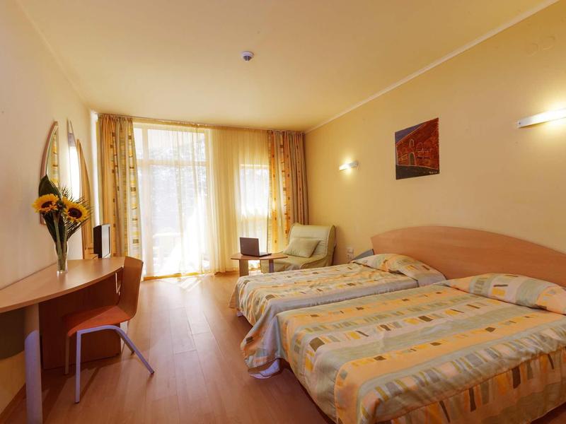 Světle hotelový pokoj se dvěma jednolůžky, psacím stolem, křeslem a velkým oknem se závěsy.