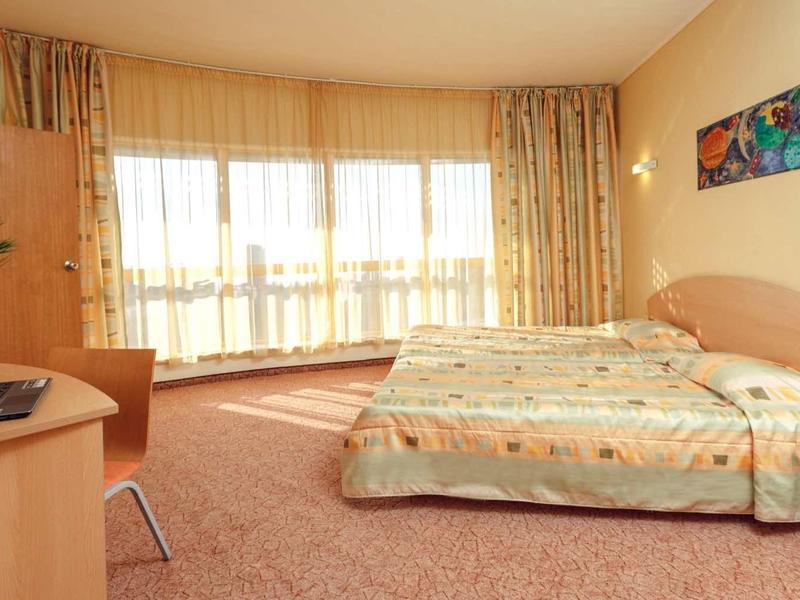 Světý hotelový pokoj s manželskou postelí, psacím stolem, židlí a velkými okny s závěsy.