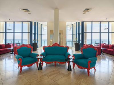 Innenraum mit blauen Sesseln und Sofas, roten Akzenten, großen Fenstern und hellem Fliesenboden.