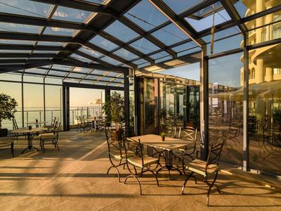 Terrazza coperta con tavoli e sedie, illuminata dal sole con vista sull'acqua in lontananza.