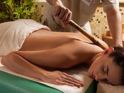 Frau liegt entspannt auf Massagetisch, erhält Rückenmassage mit Holzstab, warme Atmosphäre.