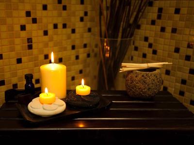 Drei brennende Kerzen auf Steinen und ein Glas mit Stäbchen vor einer gelben Fliesenwand.