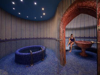 Innenraum mit blau gefliestem Boden, rundem Becken, Mauerbogen aus rotem Backstein und einer sitzenden Person.