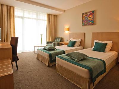 Zweibettzimmer mit beigen Wänden, zwei Einzelbetten, grün-weißer Bettwäsche und großem Fenster.