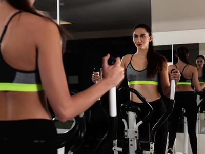 Donna in abbigliamento sportivo che si allena su una macchina da fitness davanti a uno specchio in palestra.