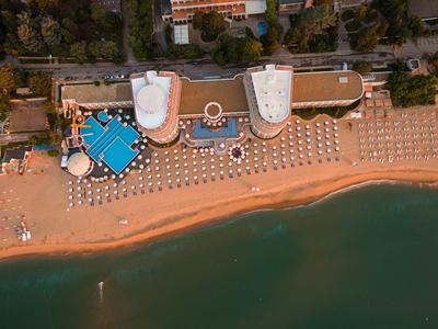 Luftaufnahme eines Strandes mit Liegestühlen, zwei weißen Gebäuden und blauem Pool am Ufer.
