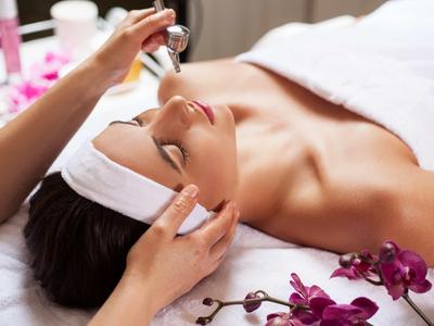 Frau liegt entspannt mit Kopfband, während Hände ein Gesichtsserum auftragen, violette Orchideen liegen daneben.