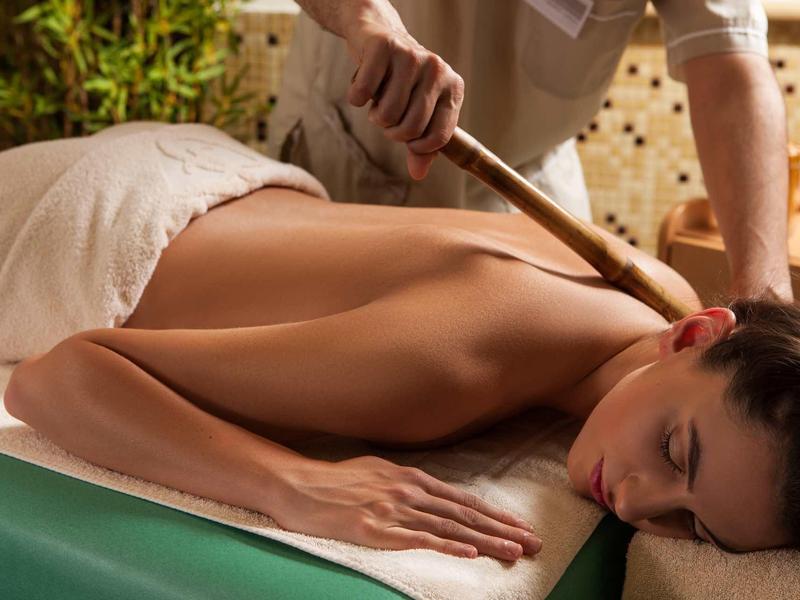 Frau liegt entspannt auf Massagetisch, erhält Rückenmassage mit Holzstab, warme Atmosphäre.