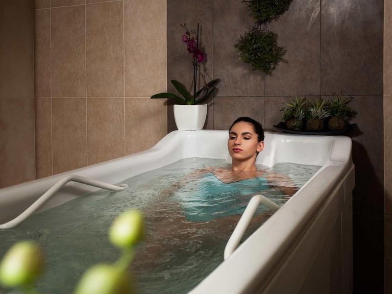 Frau entspannt in Wanne mit Handläufen, Pflanzen im Hintergrund, warmes Licht und ruhige Atmosphäre.