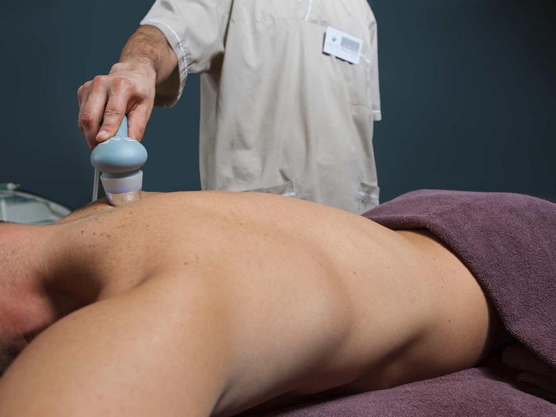 Person liegend auf Bauch, Rücken frei, wird mit Massagegerät behandelt, Hand hält Gerät über Rücken.