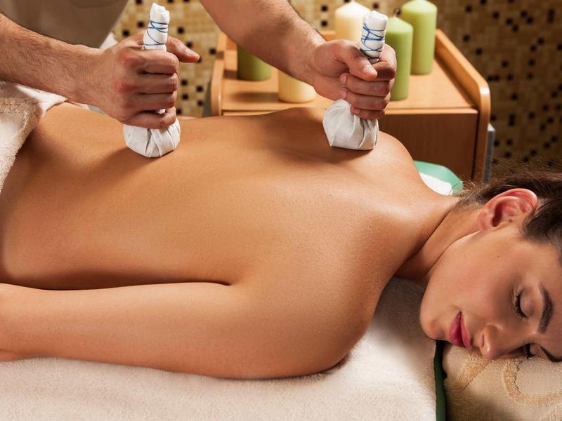 Frau liegt entspannt auf Massagebank, Rücken wird mit warmen Kräuterstempeln massiert.