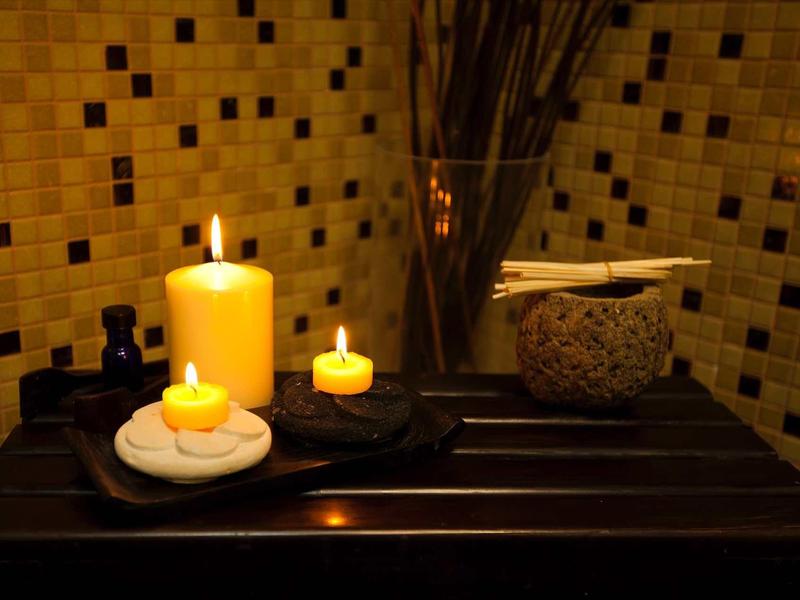 Drei brennende Kerzen auf Steinen und ein Glas mit Stäbchen vor einer gelben Fliesenwand.