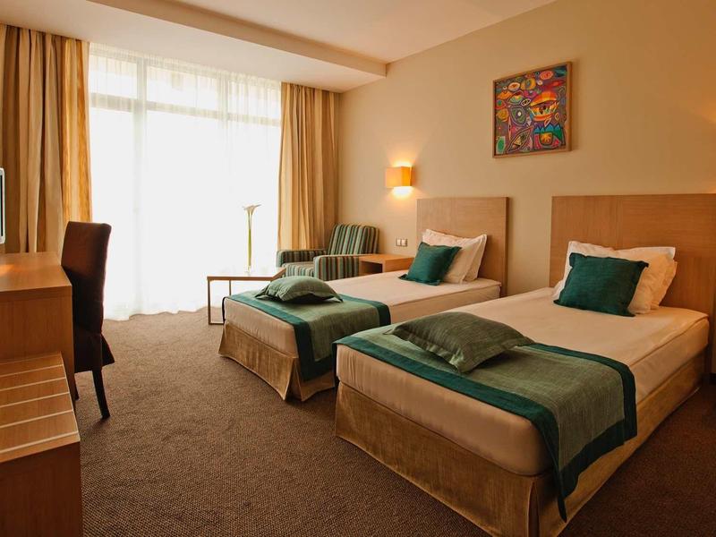 Gemütliches Hotelzimmer mit zwei Einzelbetten, Holzmöbeln und großem Fenster mit Vorhängen.