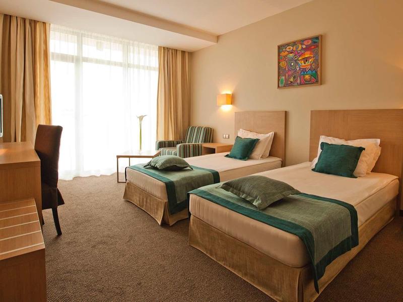 Zweibettzimmer mit beigen Wänden, zwei Einzelbetten, grün-weißer Bettwäsche und großem Fenster.