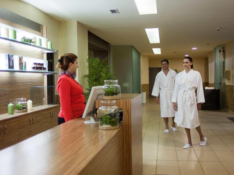 Rezeptionistin begrüßt zwei Gäste im Bademantel in einem modernen Wellnessbereich.