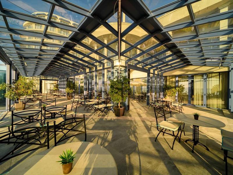 Moderne Terrasse mit Glasdach, mehreren Tischen und Stühlen, sowie Sonnenschein und Pflanzen.