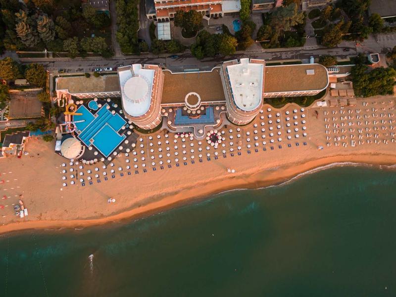 Luftaufnahme eines Strandes mit Liegestühlen, zwei weißen Gebäuden und blauem Pool am Ufer.