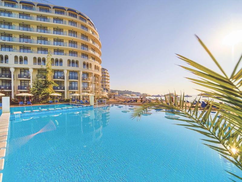 Großer Hotelpool mit klar blauem Wasser, Sonnenschirmen und einem mehrstöckigen Gebäude im Hintergrund.
