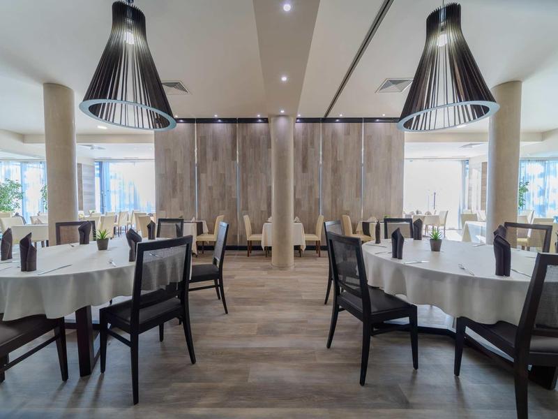 Moderner Restaurantraum mit Holzfußboden, runden Tischen mit weißen Tischtüchern und großen Lampen.