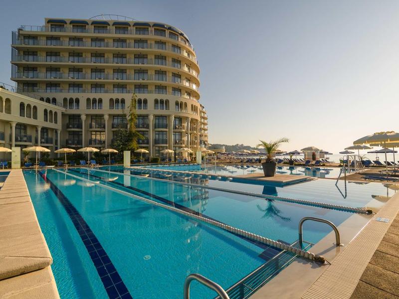 Moderner Hotelkomplex neben großem Außenpool mit klar blauem Wasser und Liegestühlen.