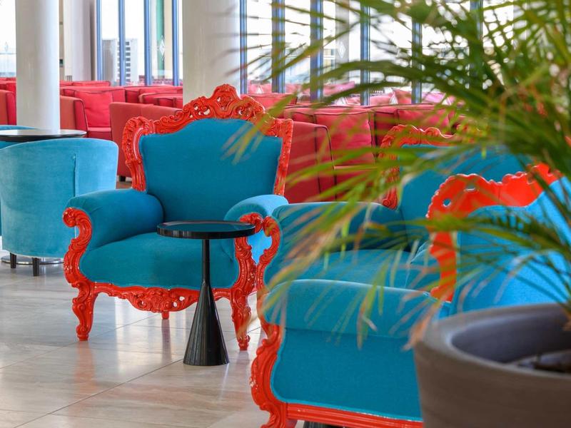 Helle Lounge mit blauen Sesseln und roten, verzierten Holzrahmen, Pflanzen im Vordergrund.