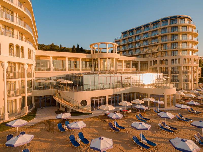 Modernes Hotel mit großen Glasfenstern, Sonnenschirmen und Liegestühlen an einem hellen Tag.