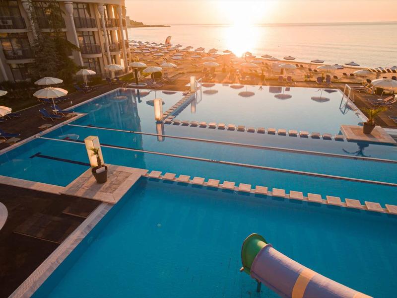 Großer Hotelpool mit Wasserrutsche bei Sonnenuntergang am Meer, Liegestühle und Sonnenschirme rundherum.