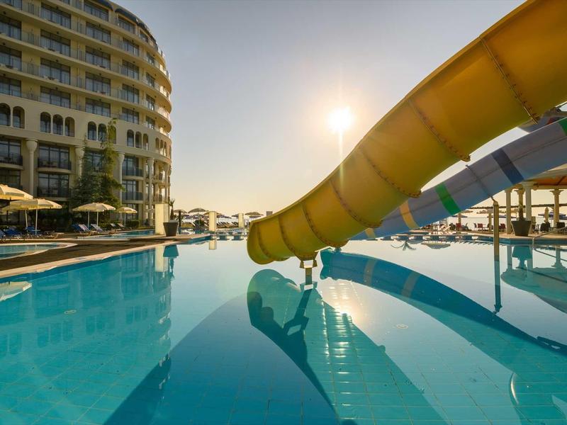 Großer Pool mit gelber Wasserrutsche, Sonnenschirmen und rundem Hotelgebäude bei Sonnenlicht.