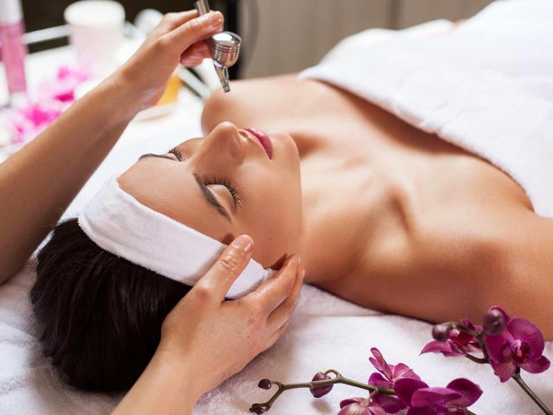 Frau liegt entspannt mit Kopfband, während Hände ein Gesichtsserum auftragen, violette Orchideen liegen daneben.