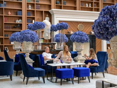 Elegante zona de salón de hotel con sillones azules, arreglos florales y una pared de libros al fondo.