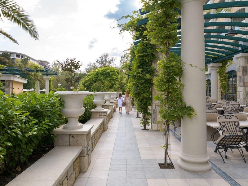 Amplia terraza con columnas y área de asientos junto a plantas verdes y palmeras bajo cielo nublado.