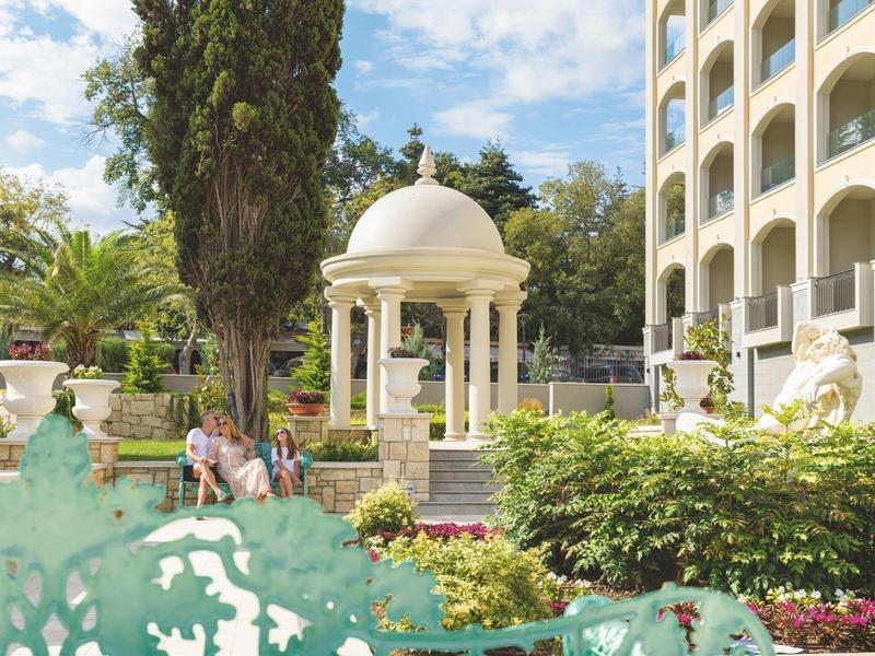 Jardín con pabellón blanco, rodeado de áreas verdes y un edificio de varios pisos al fondo.