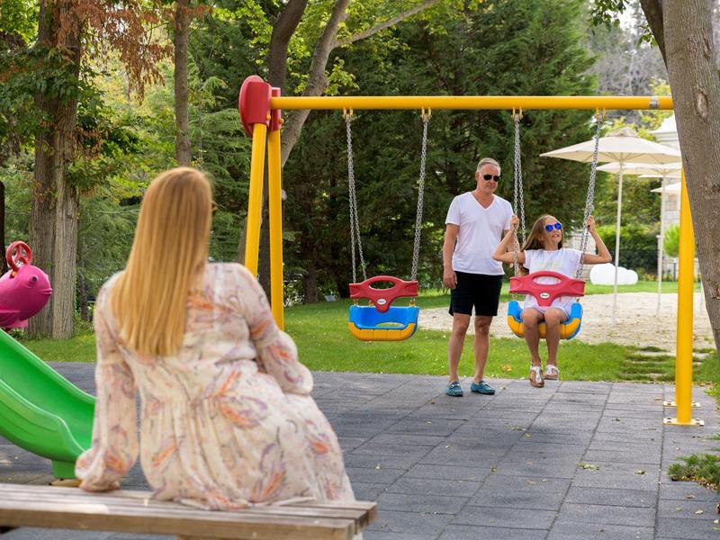 Mujer en un banco observa a dos niños en un columpio en el parque con clima soleado.