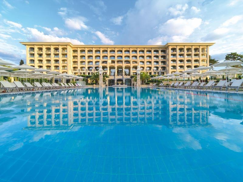 Gran hotel de varios pisos con una gran piscina en primer plano bajo un cielo azul.