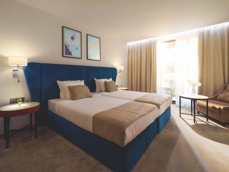 Habitación de hotel moderna con cama grande, mesas de noche, sillones y luz brillante a través de cortinas.