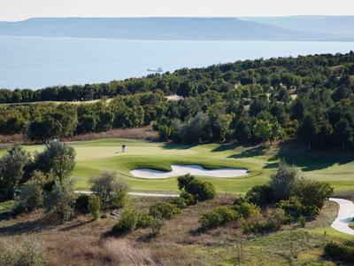 Zelené golfové hřiště s pískovými bunkry v kopcovité krajině a mořem v pozadí.