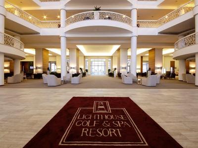Große Hotel-Lobby mit hohen Decken, weißen Säulen, Sesseln und rotem Teppich mit Resort-Logo.