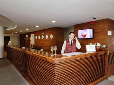 Hotelová recepce s recepčním u moderní dřevěné přepážky a dekorativními zrcadly na stěně.