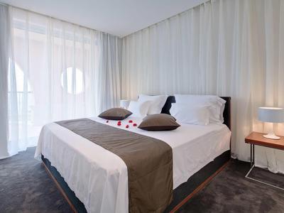 Moderní hotelový pokoj s velkou postelí, hnědými akcenty a denním světlem skrz závěsy.