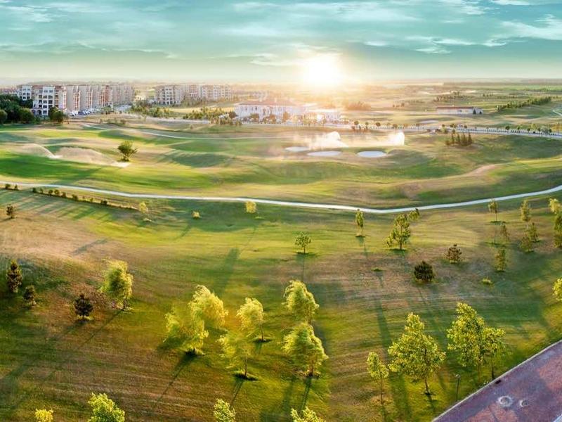 Panoramatický pohled na rozsáhlé golfové hřiště se zelenými trávníky, stromy a slunečnou oblohou.