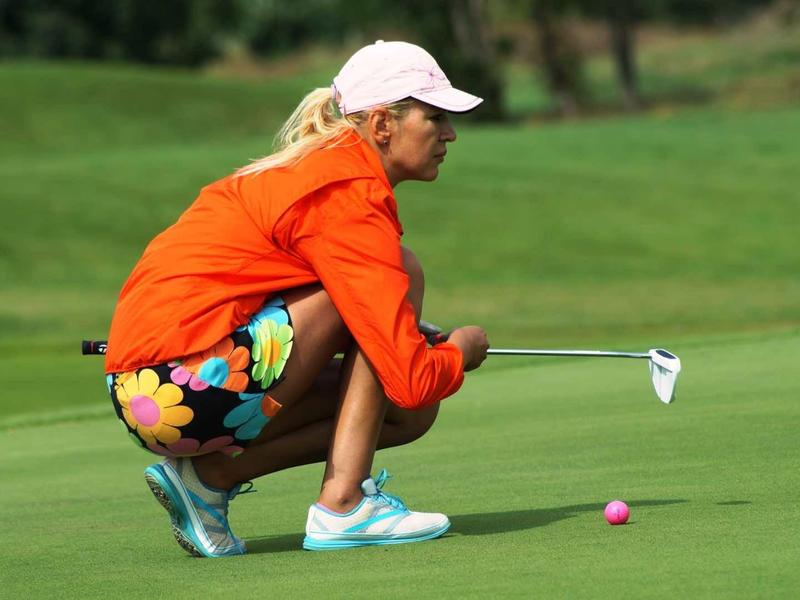 Žena v oranžovém svetru a barevných kalhotách klečí na golfovém hřišti a míří na golfový míček.