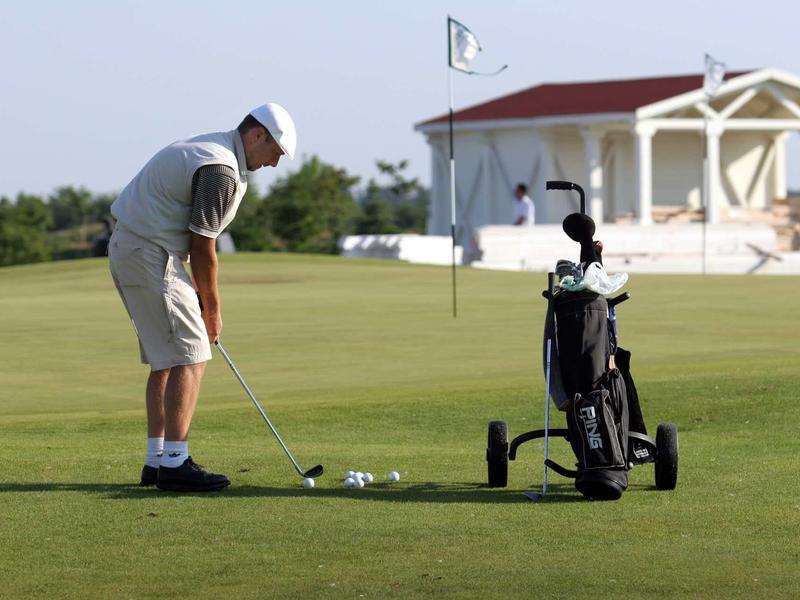 Muž hrající golf u golfového vozíku na zeleném golfovém hřišti s klubovnou v pozadí