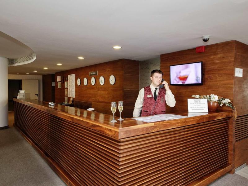 Hotelová recepce s recepčním u moderní dřevěné přepážky a dekorativními zrcadly na stěně.