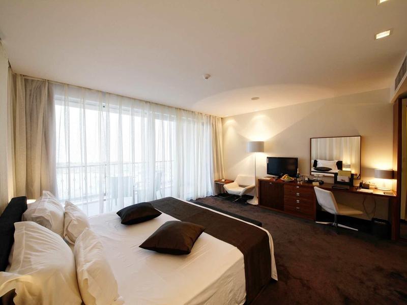 Moderní hotelový pokoj s dvoulůžkem, balkonem a psacím stolem za denního světla.