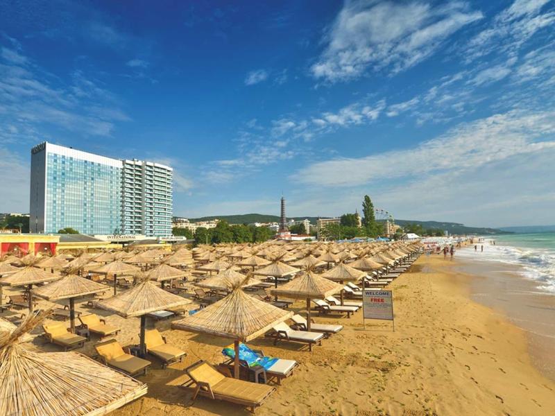 Large plage de sable avec parasols, transats et hôtels en arrière-plan sous un ciel clair.