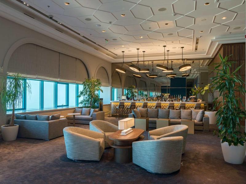 Espace lounge d'hôtel moderne avec fauteuils confortables, tables et plantes près de grandes fenêtres.